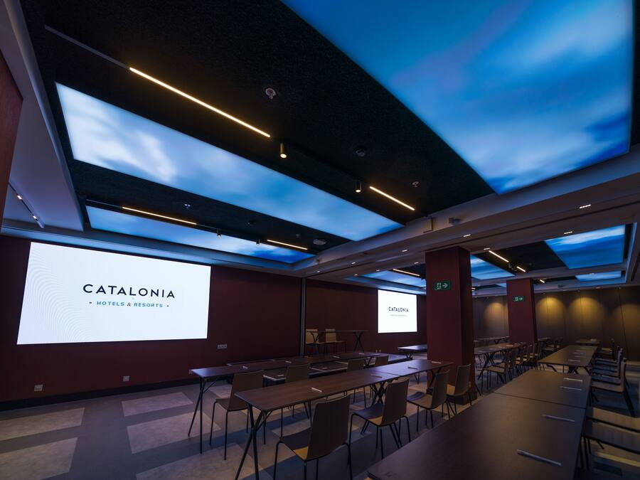 Una sala de conferencias con una pantalla grande que muestra 'CATALONIA HOTELS & RESORTS', mesas, sillas y un techo con diseño que simula el cielo.