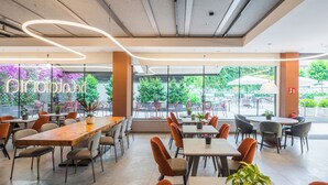 Un café moderno con mesas de madera y sillas naranjas, ventanas amplias y plantas en macetas.