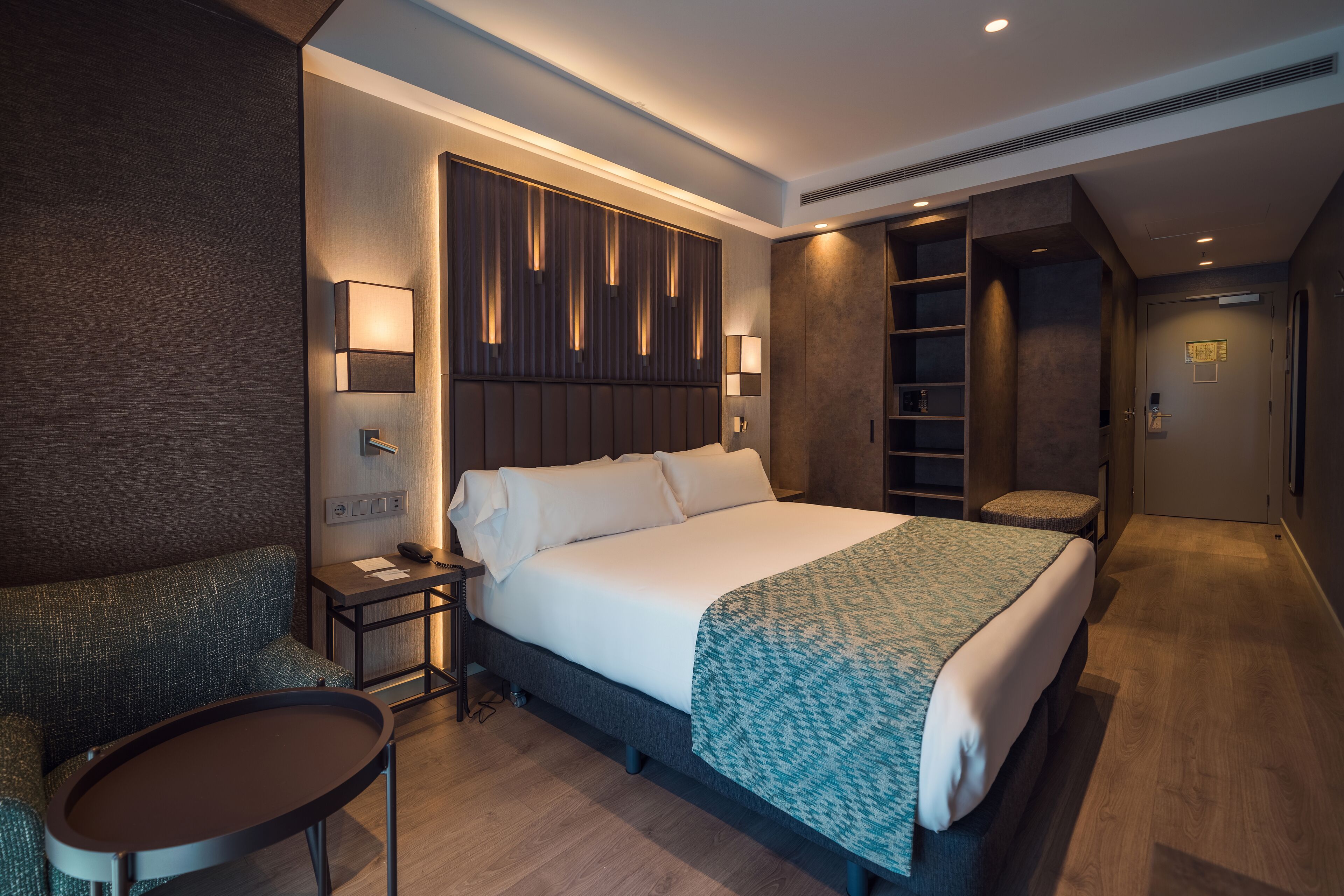 Ein modernes Hotelzimmer mit einem großen Bett, einer Sitzecke mit kleinem Tisch, einem Schreibtisch mit Lampe und einem Kleiderschrank mit Einbauschränken.