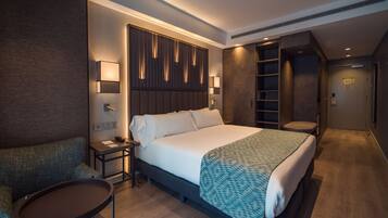 Ein modernes Hotelzimmer mit einem großen Bett, einer Sitzecke mit kleinem Tisch, einem Schreibtisch mit Lampe und einem Kleiderschrank mit Einbauschränken.