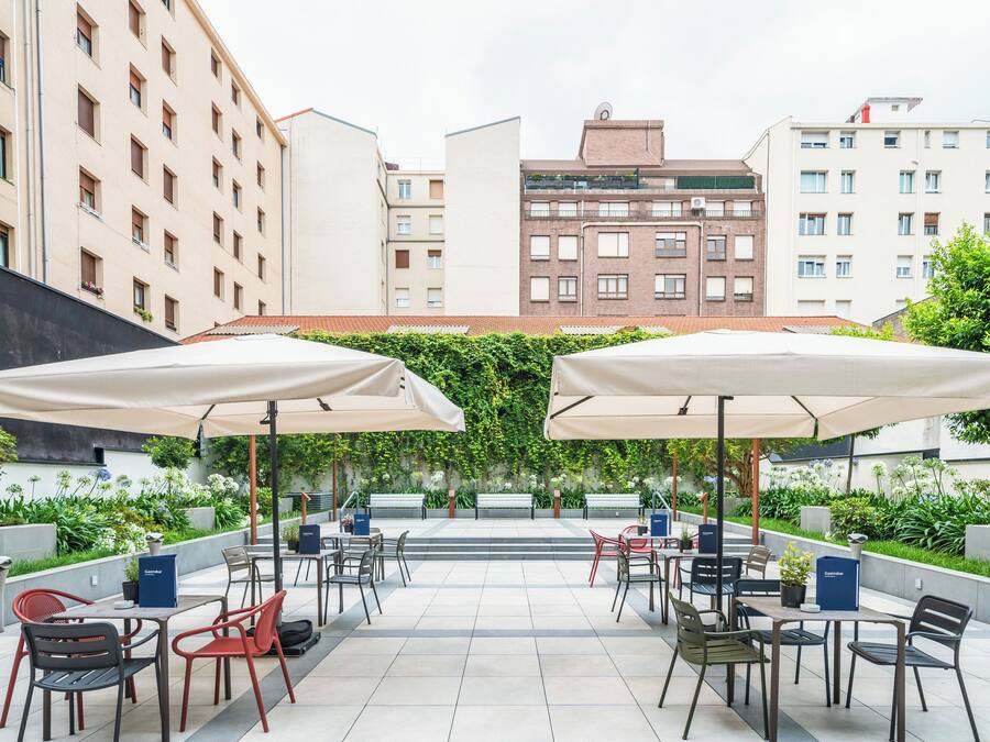 Une terrasse extérieure avec des tables et des chaises, protégée par des parasols et entourée de verdure et de bâtiments.
