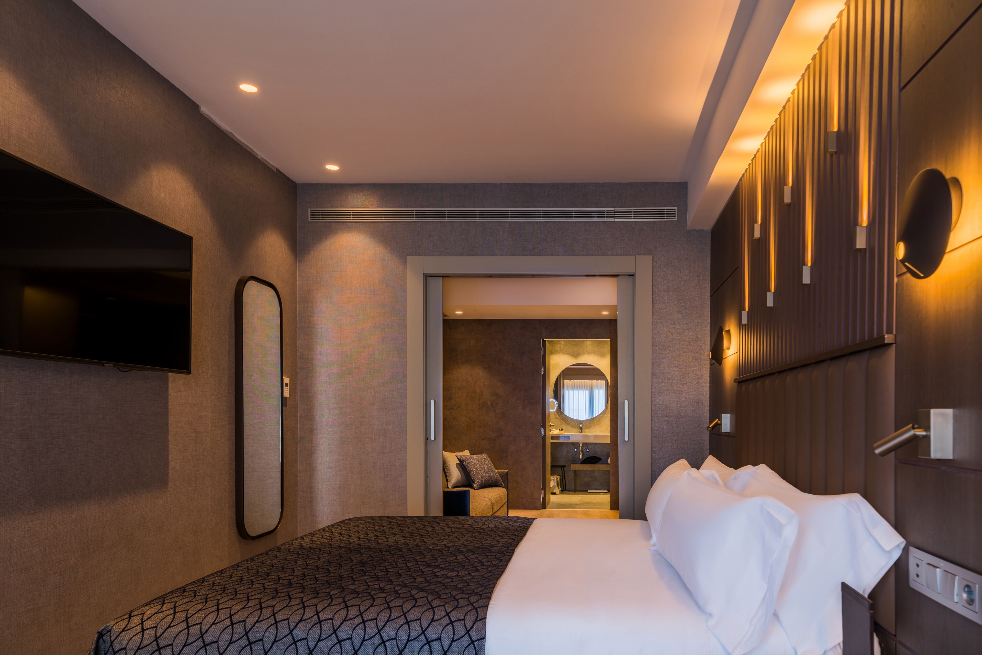 A modern hotel room with a large bed, a flat-screen TV, a mirror, and a bathroom visible through an open door.