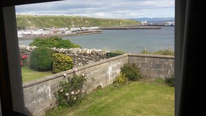 A view from a window overlooking a coastal area with a grassy lawn, a stone wall, and a body of water. - Bungalow with sea-view, near small town of Thurso.  (Thurso)