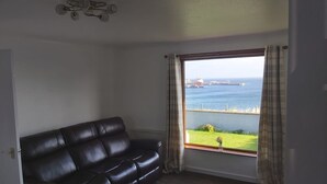 A room with a black leather sofa, a view of the sea, and a window with curtains. - Bungalow with sea-view, near small town of Thurso.  (Thurso)
