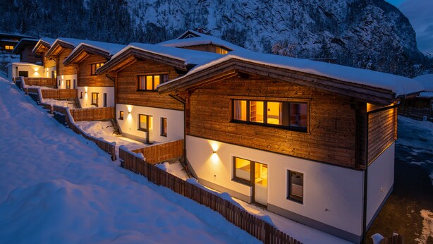 A row of wooden cabins with snow-covered roofs, set against a mountainous backdrop. - Chalet houses (Oetz)