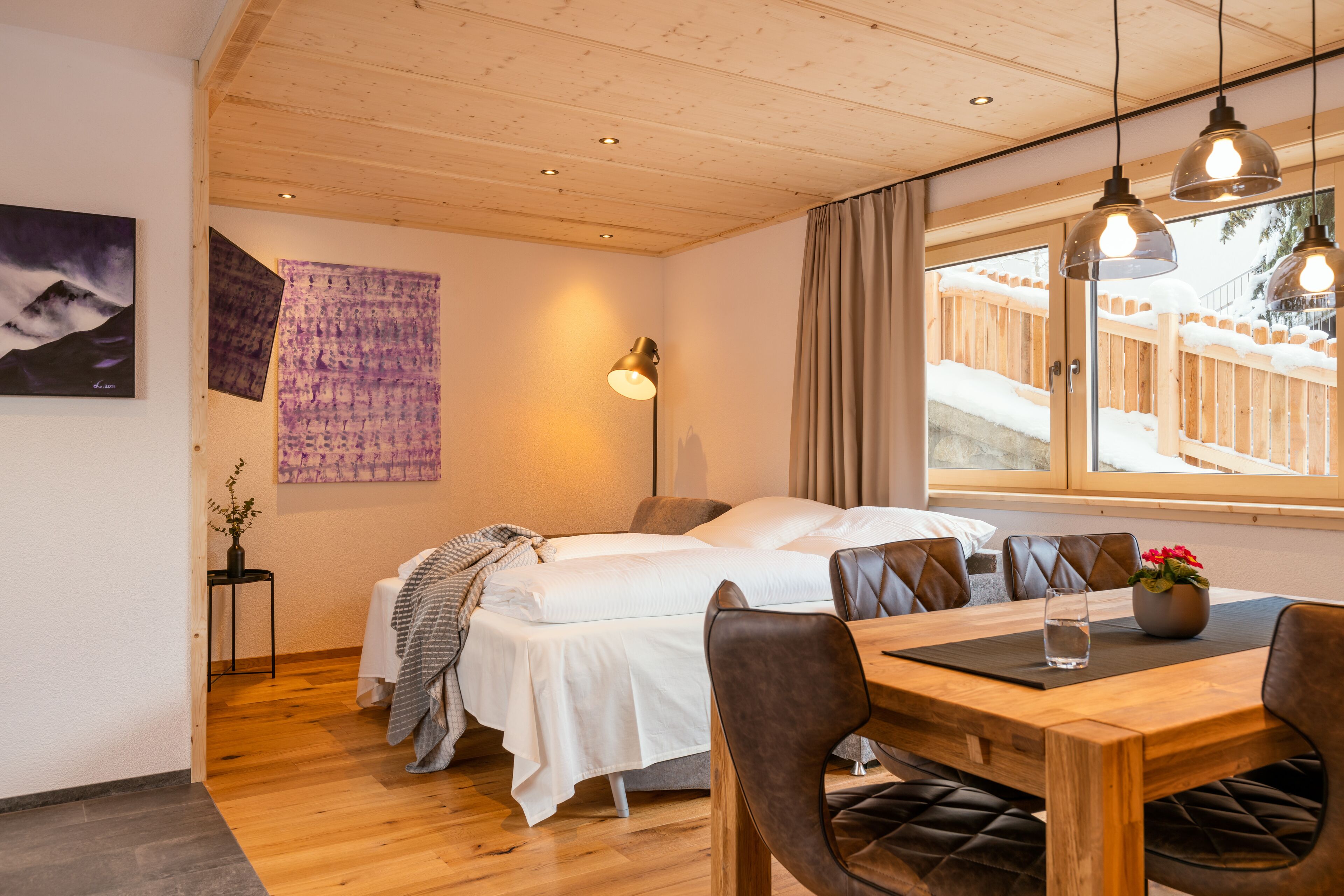 Ein gemütliches Zimmer mit einem Bett, einem Esstisch mit Stühlen, einer Vase mit Blumen und einem Fenster mit Blick auf einen Holzzaun.