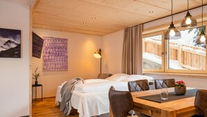 A cozy room with a bed, a dining table with chairs, a vase with flowers, and a window with a view of a wooden fence. - Chalet houses (Oetz)
