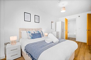 A modern bedroom with a large bed, bedside tables, a nightstand, and a bathroom visible through an open door. - Luxury One Bed Apartment - JQ (West Midlands)