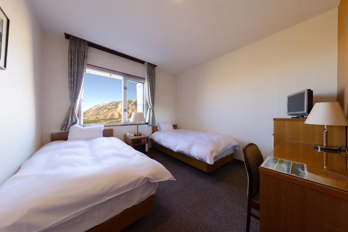 Habitación de hotel con dos camas, un escritorio con lámpara, televisión y vistas a las montañas desde la ventana.