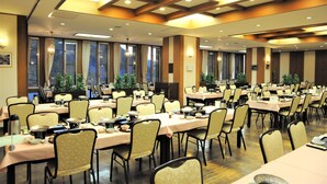 A large banquet hall with multiple tables and chairs, arranged in rows, with large windows and ceiling lights.