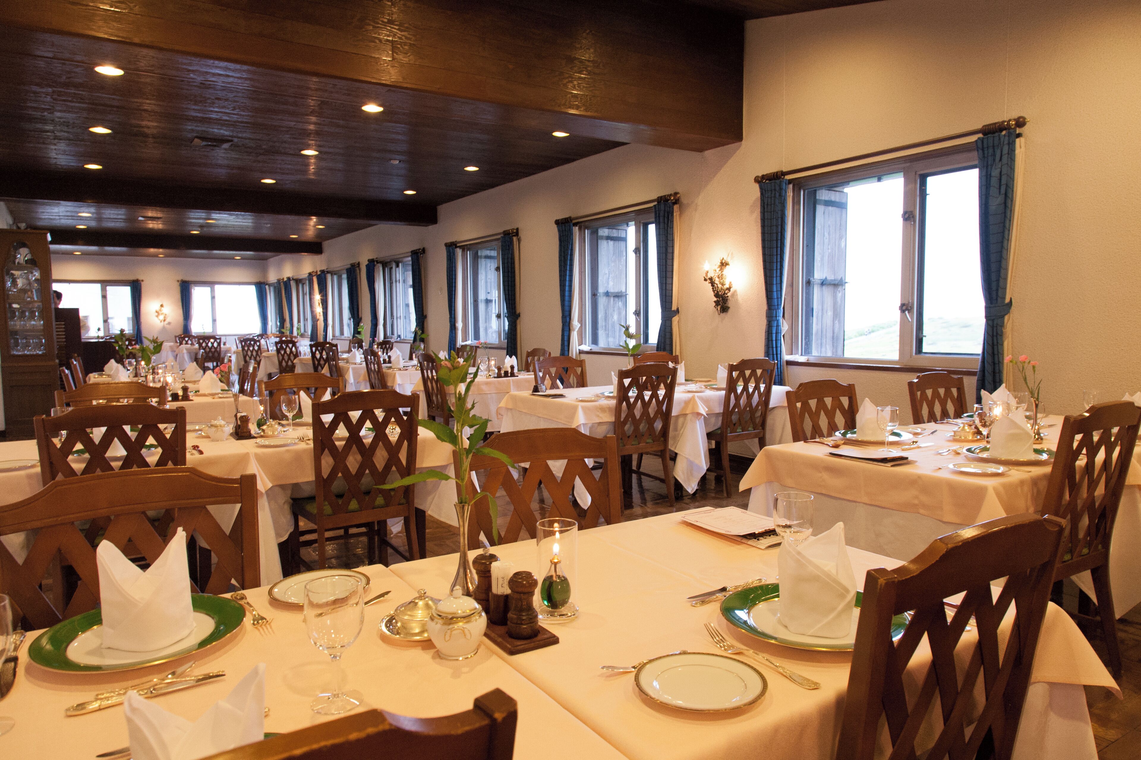 Un restaurante con varias mesas dispuestas para comer, sillas de madera y ventanas amplias con cortinas.