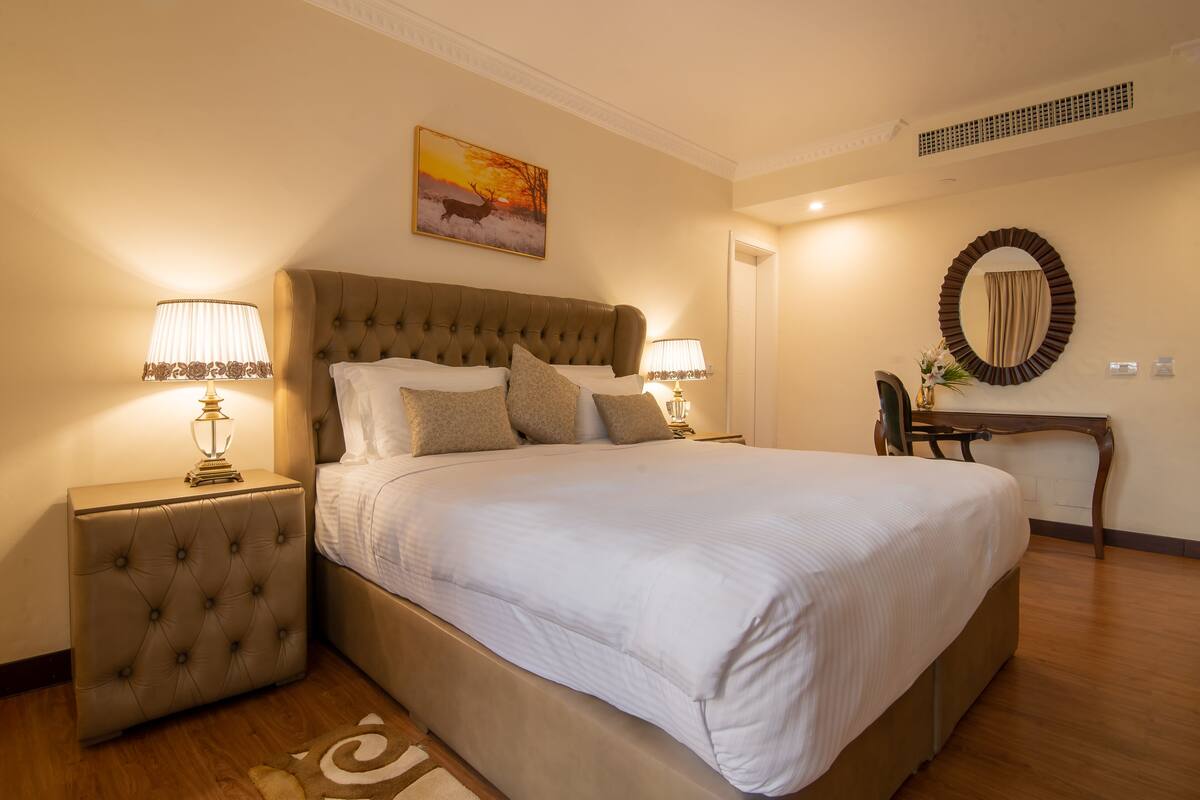 Une chambre spacieuse avec un grand lit, deux tables de chevet avec des lampes, un miroir rond et un tableau au mur.