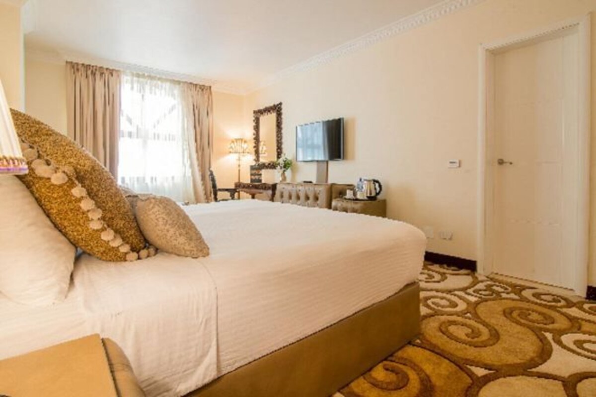 A hotel room with a large bed, a television, a desk with a lamp, and a patterned carpet.