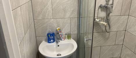 Un baño con lavamanos pedestal, cabina de ducha con mampara de vidrio y artículos de aseo sobre el lavamanos.