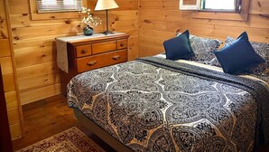 A wooden cabin bedroom with a patterned bedspread, a bedside table with a lamp, and a window with blinds. - Cottage by the Lake #3 - Blossoms (Bristol)