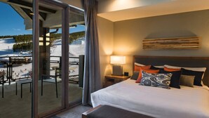 A hotel room with a bed, a nightstand, a lamp, a view of a snowy landscape through a glass door, and a balcony with chairs. - Grand Colorado Peak 8 (Breckenridge)