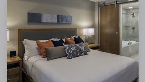 A neatly made bed with multiple pillows, a bedside lamp, and a nightstand with a device. There is a wooden wardrobe and a glass shower area visible. - Grand Colorado Peak 8 (Breckenridge)