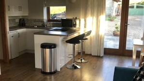 A compact kitchen with a central island, two bar stools, a stainless steel trash bin, and a view of the outside through a window. - Rural Retreat in Warwickshire (Rowington)