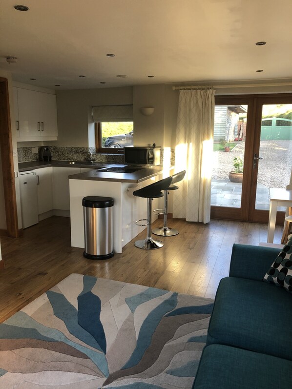 A compact kitchen with a central island, two bar stools, a stainless steel trash bin, and a view of the outside through a window. - Rural Retreat in Warwickshire (Rowington)