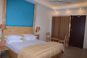 A bedroom with a bed, two bedside lamps, a wooden nightstand, a table with chairs, and a brown door. - Coconhuts Beach Resort-neil Island (Port Blair)