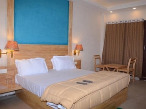 A hotel room with a large bed, two bedside lamps, a wooden headboard, a small table with a chair, and a curtain.
