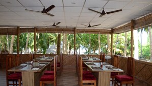 A restaurant with long wooden tables and red cushioned chairs, large windows with a view of palm trees, and ceiling fans. - Coconhuts Beach Resort-neil Island (Port Blair)