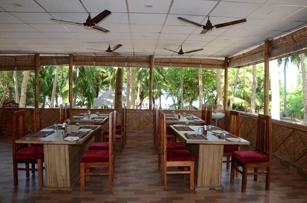 Un restaurante con largas mesas de madera y sillas con acolchado rojo, amplias ventanas con vistas a palmeras y ventiladores de techo.