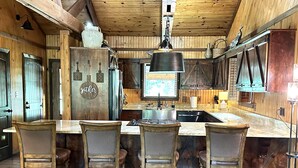 A rustic kitchen with wooden walls, a central island with bar stools, and a hanging light fixture. - Waterfront 4 bedroom Cabin with Outdoor Amenities (Broken Bow)