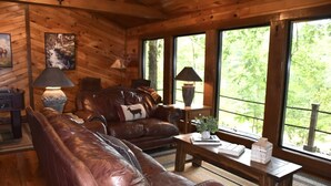 A cozy cabin interior with leather furniture, a fireplace, and large windows offering a view of the outdoors. - Waterfront 4 bedroom Cabin with Outdoor Amenities (Broken Bow)