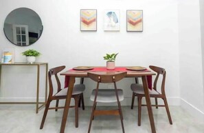 A dining area with a round table, four chairs, a small side table with a plant, and framed artwork on the wall.