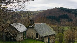 A rustic stone building with a sloped roof, situated on a grassy hillside with a forested backdrop.