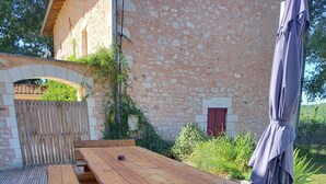 Une table de pique-nique en bois, avec des bancs, installée sur une terrasse en bois, entourée de verdure et d’un bâtiment en pierre.