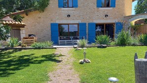 Une maison en pierre à deux étages, avec des volets bleus, un chemin de gravier menant à l’entrée et un petit jardin agrémenté de plantes et d’une clôture en bois.