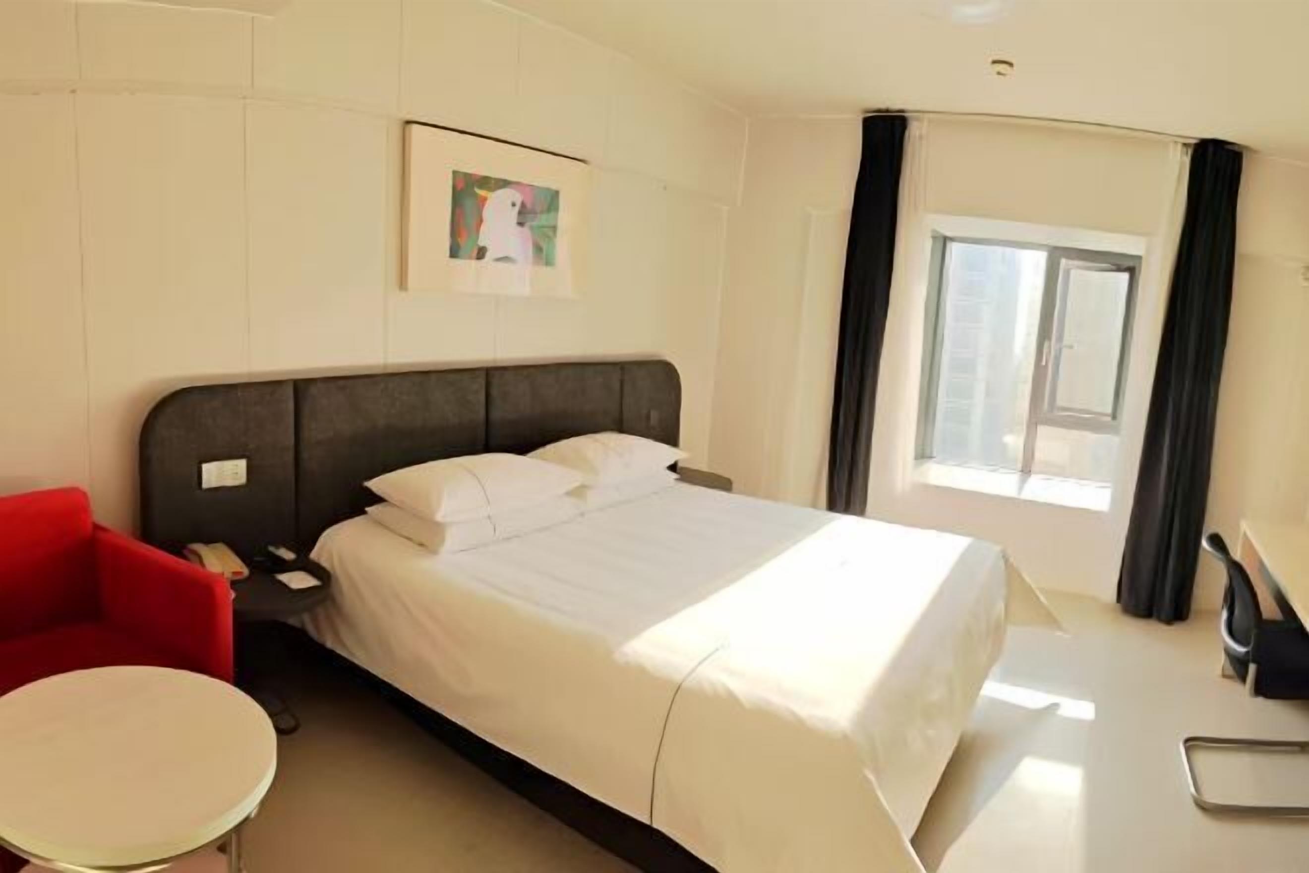 A hotel room with a bed, a red chair, a small round table, a desk, and a window with curtains.