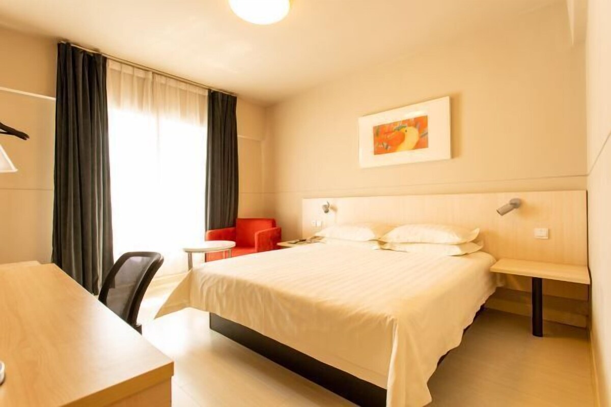 Une chambre d’hôtel avec un grand lit, un bureau, une chaise et une petite table. Il y a une fenêtre avec des rideaux, un tableau au mur et une lampe sur la table de chevet.