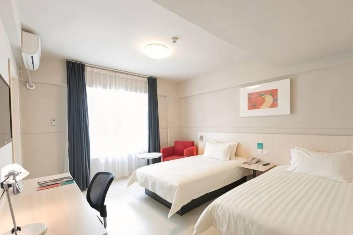 Quarto de hotel com duas camas, uma mesa, uma cadeira, uma mesinha e uma janela com cortinas.