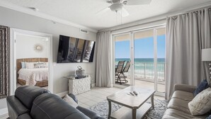 A spacious living room with a ceiling fan, a flat-screen TV, a balcony with a view of the ocean, and a cozy seating area.