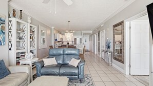 A spacious living room with a blue leather sofa, a beige armchair, and a coffee table. There is a chandelier, a ceiling fan, and a large mirror on the wall.