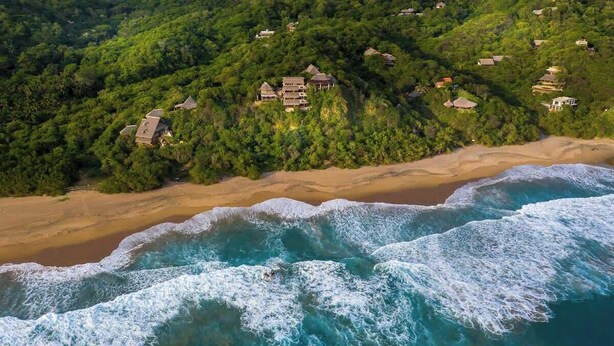 An aerial view of a coastal area with several houses surrounded by lush greenery, a sandy beach, and waves crashing onto the shore. - Lazuli Cielo - Beachfront Loft (Santa María Tonameca)