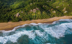 Vista aérea de una zona costera con varias casas rodeadas de exuberante vegetación, una playa de arena y olas rompiendo en la orilla.