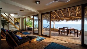 A spacious living area with a large sliding glass door leading to a balcony with a view of the ocean. There is a wooden staircase on the left, a sofa with cushions, and a dining table with chairs on the balcony. - Lazuli Cielo - Beachfront Loft (Santa María Tonameca)