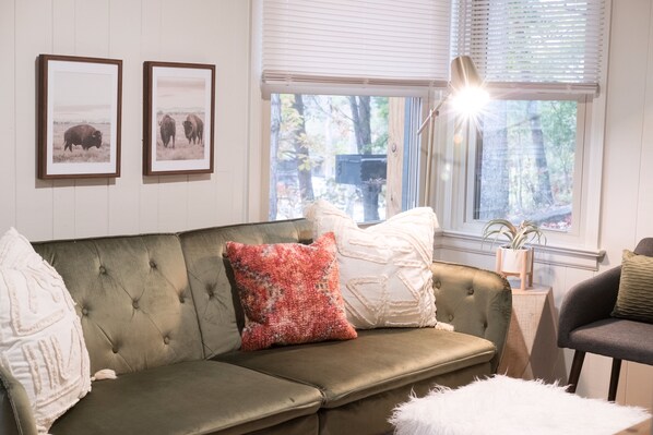 A living room with a green sofa, two framed pictures on the wall, and a window with blinds. - Minimalist-Style Apartment within walking distance of Downtown Johnson City (Johnson City)