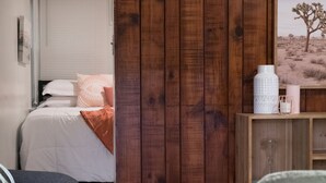 Un dormitorio con un armario de madera, una cama con edredón blanco y manta naranja, y una mesita de noche con un jarrón blanco y una vela.