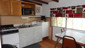 A compact kitchen with white cabinets, a microwave, a stove, and a coffee maker. There's a window with a view of trees and a table with a chair.