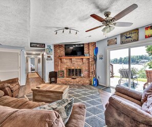 A spacious living room with a brick fireplace, a ceiling fan, and a large TV mounted above it.