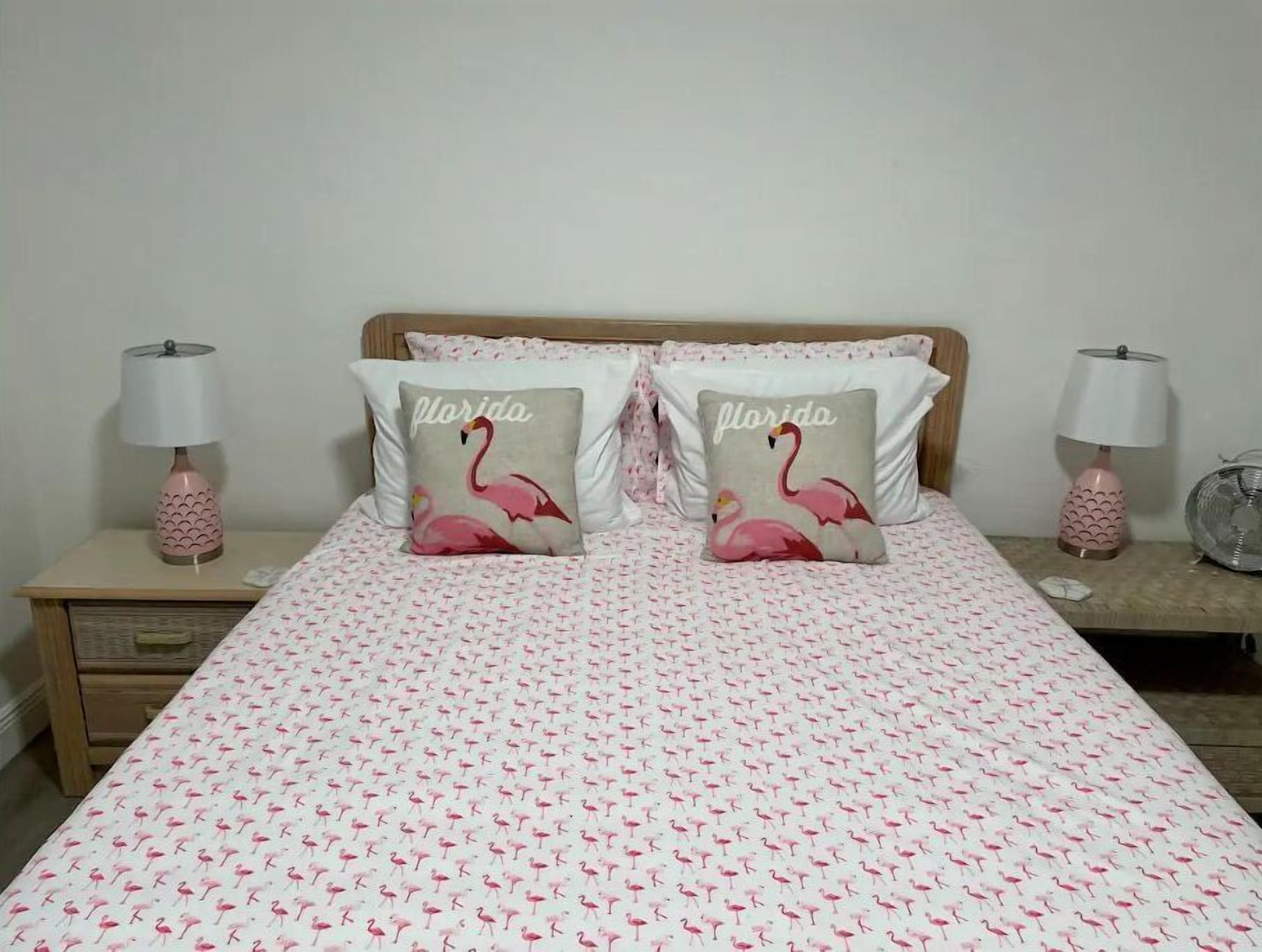 Un letto con copriletto a motivi rosa e bianchi di fenicotteri, due cuscini con disegni di fenicotteri e lampade abbinate su entrambi i lati.