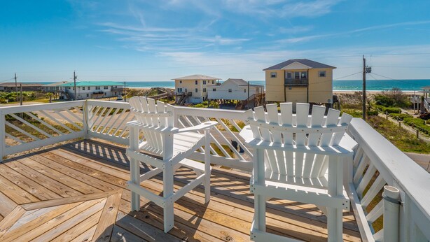 Eine Holzterrasse mit weißen Stühlen mit Blick auf einen Strand, im Hintergrund Häuser.