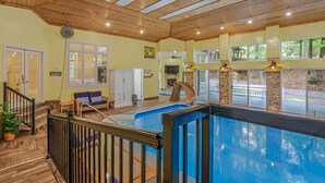 A spacious indoor swimming pool with a wooden deck and a slide, surrounded by large windows and a stone wall.