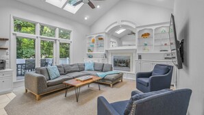 A spacious living room with a sectional sofa, a coffee table, and a fireplace. There are large windows and a ceiling fan.