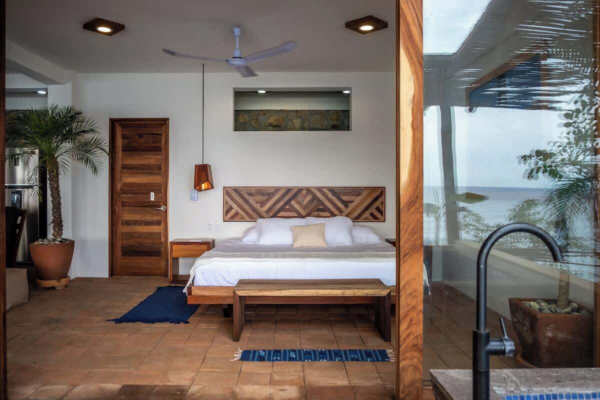 Un dormitorio con cama, cabecera de madera, ventilador de techo y vista al océano a través de un ventanal.